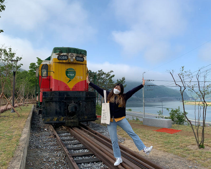 creatrip editor posing on abandoned train track with namhan river, and red yellow green train car in background in danyang south korea