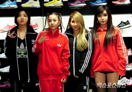 K-POPグループ2NE1のメンバーたちが、赤と黒のAdidasジャケットを着て、ショップの背景の前に立っている。