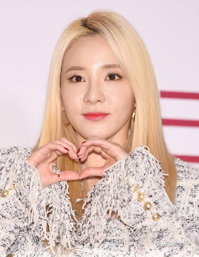 歌手パク・サンダラ、ハートのポーズで手を作りながらカメラに向かって微笑んでいる。彼女は白いフリンジのついた服を着ている。