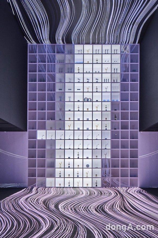  HYBE 음악 환상 트로피 전시 공간, 동상들이 전면에 있고 뒤에는 긴 얇은 빛의 선들이 있음.