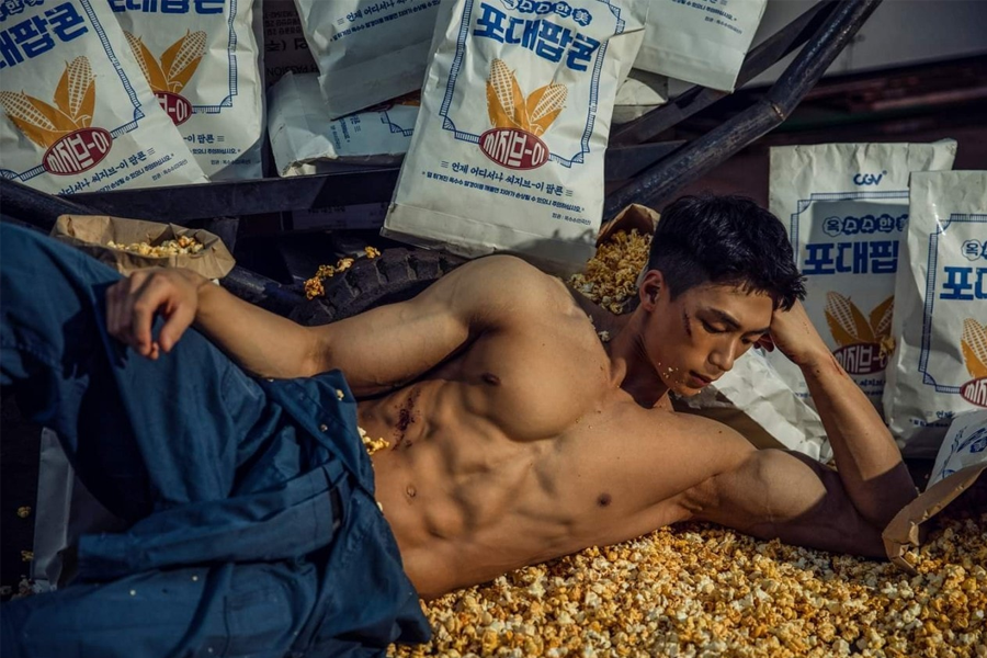 一位上身赤裸的肌肉男士躺在散落的爆米花上，背景是多个标有韩文的爆米花大包装袋。