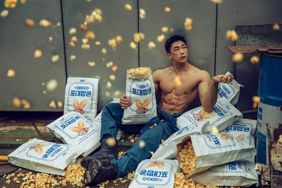 一位上身赤裸的男士坐在爆米花堆中，持有大包的爆米花袋，周围是散落的爆米花。