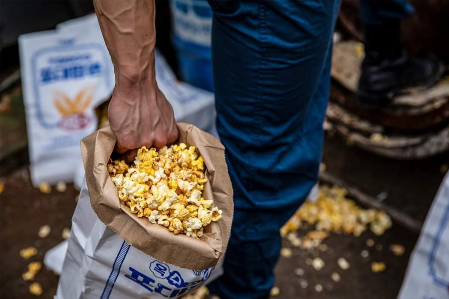 一只手抓着一袋装满爆米花的纸袋，背景模糊有多个标有韩文的爆米花包装。