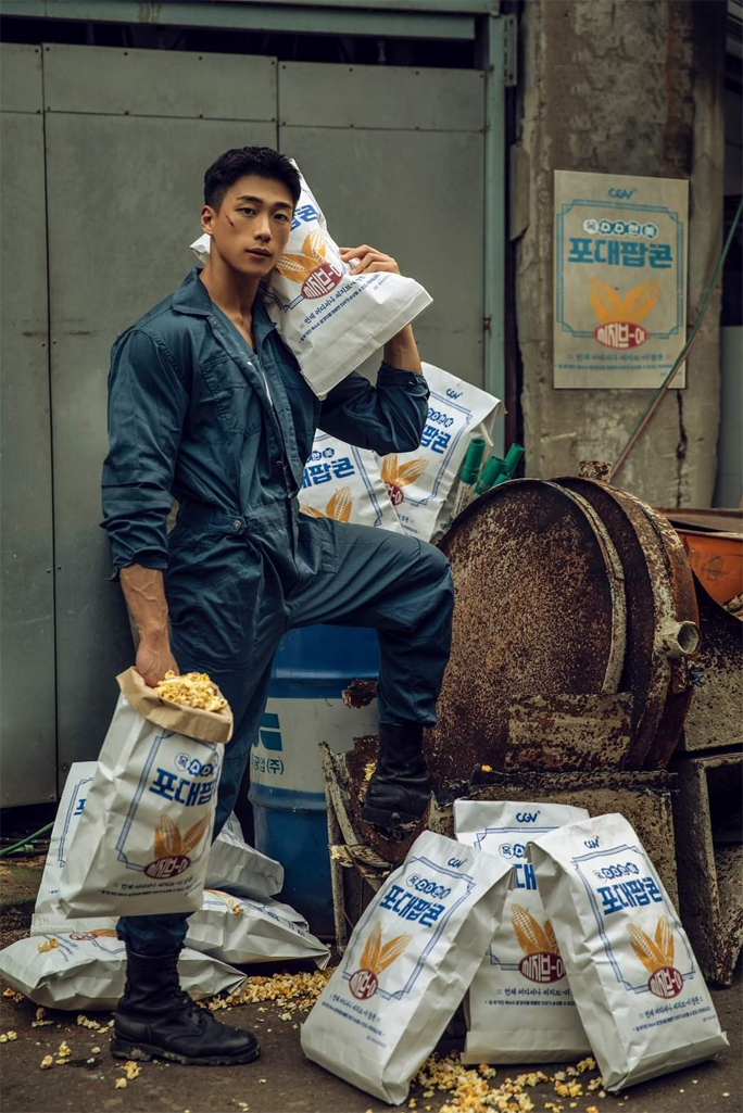 一位穿着蓝色连体服的男士，站在一个工业背景中，手提和肩扛大袋爆米花，周围是多个爆米花袋。