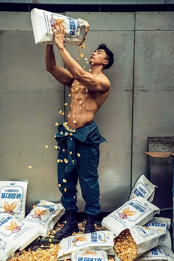 一位上身赤裸的肌肉男士用大量爆米花倾洒在身上，脚下散落着多个标有韩文的爆米花大包装袋。