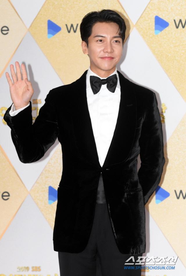 スーツを着た韓国の男性セレブがイベントに参加している姿。彼は手を振って挨拶しており、背景にはイベントのロゴが見える。