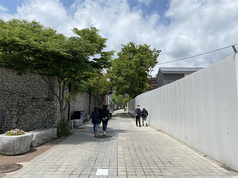 A peaceful alleyway lined with trees and traditional walls, leading towards Bukchon Hanok Village, with people strolling on a sunny day in Seoul.