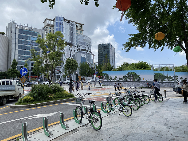 A bikeway filled with green rental bicycles and people walking along a modern street with buildings and trees, located near a large intersection in Seoul.