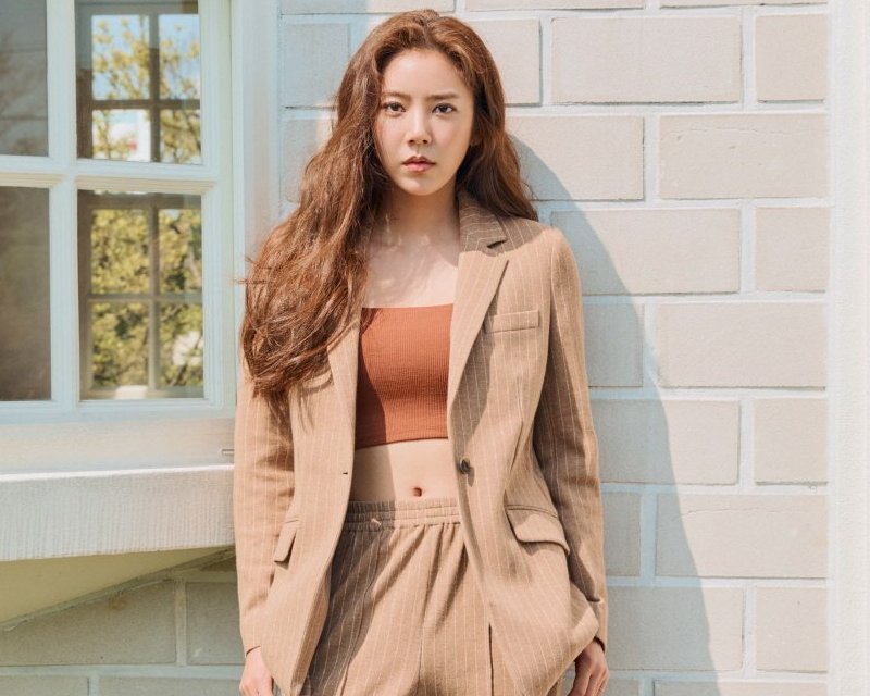 韓國藝人孫淡妃的照片，身穿棕色西裝和短上衣，站在淺色磚牆前。