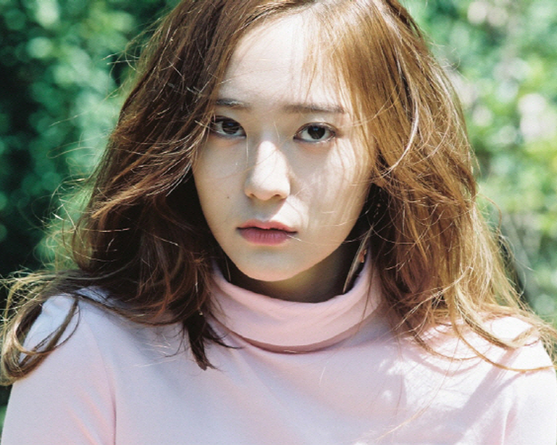 韓國藝人Krystal的照片，穿著粉紅色襯衣，背景為綠色植物。