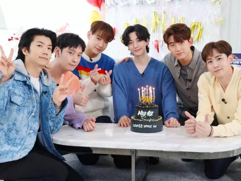 สมาชิกวง 2PM นั่งรอบโต๊ะเค้กฉลองวันเกิด บรรยากาศเต็มไปด้วยลูกโป่งและคำว่า 'HAPPY BIRTHDAY' ในพื้นหลัง