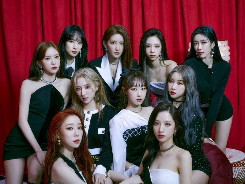 สมาชิกวง Cosmic Girls นั่งอยู่หน้าฉากพื้นหลังสีแดงสวยงาม ทุกคนใส่ชุดแฟชั่นล้ำสมัย