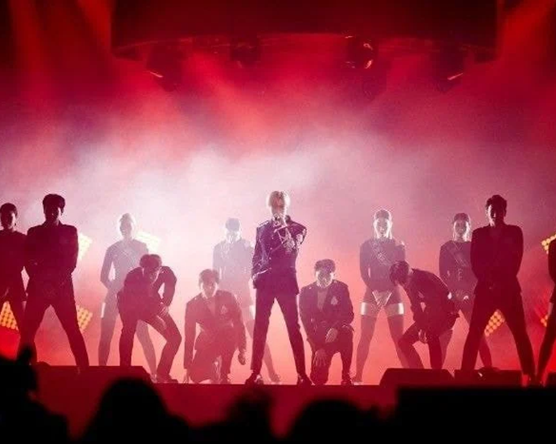K-Pop-Idol auf der Bühne bei einem Konzert mit Tänzern, beleuchtet von roten Lichtern.