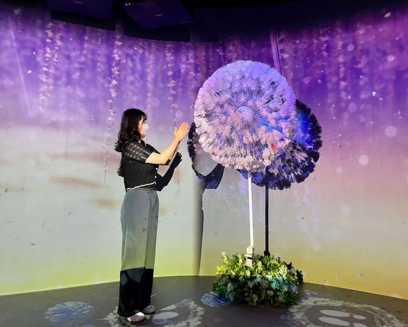 Giovane visitatrice accanto a un grande dente di leone alla mostra Flowers By Naked Hongdae.