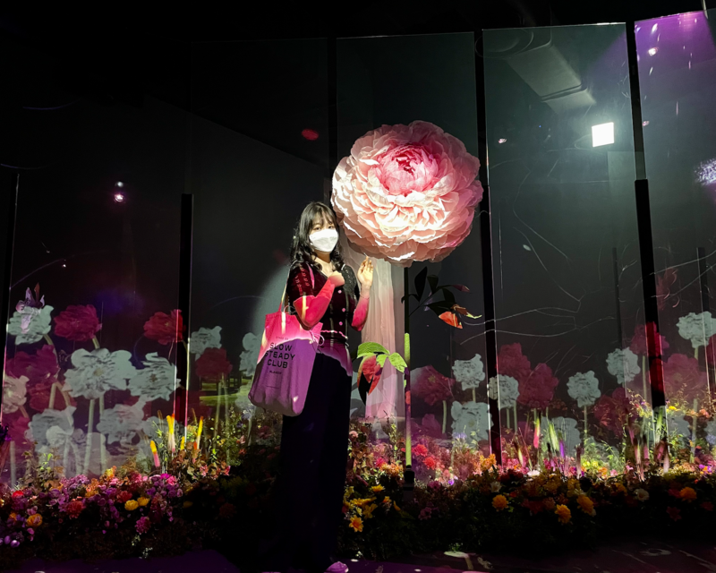 Visitatrice posa accanto a un grande fiore rosa nella zona Grande Giardino Fiorito di Flowers By Naked.