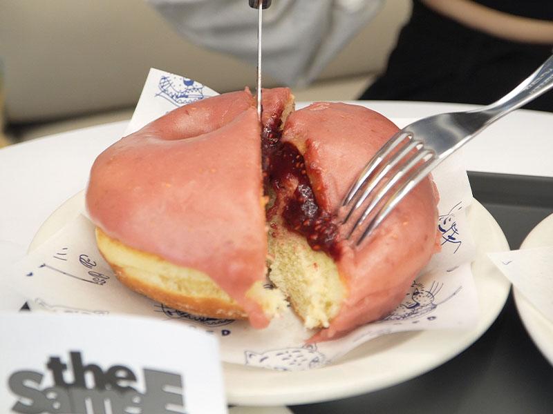 一個覆滿粉色糖霜嘅拉斯比利果醬甜甜圈，正喺用刀叉進行切割。