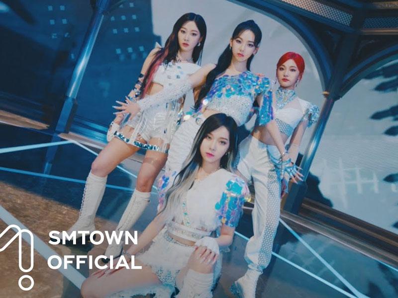 สมาชิกวง aespa ในชุดสีขาว พร้อมบรรยากาศการแสดงเพลง "Next Level" บนเวทีที่มีสิ่งก่อสร้างทันสมัย