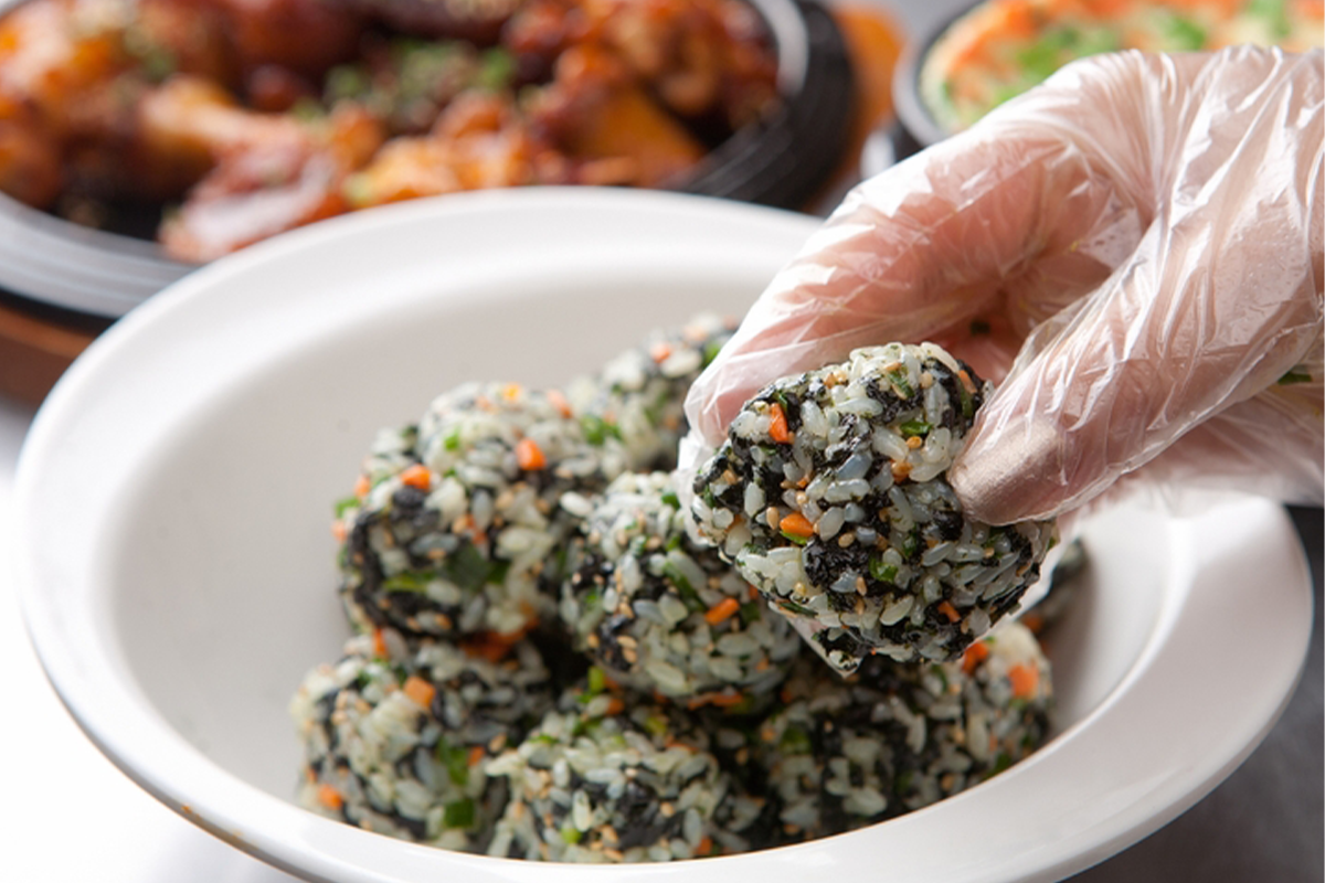 這張圖片顯示一雙帶著手套的手，正在抓一粒韓國飯團，這種飯團通常是用紫菜和米飯混合製成的，飯粒中還有蔬菜，這是一種常見的韓式食物，尤其是用來解辣的食物之一。