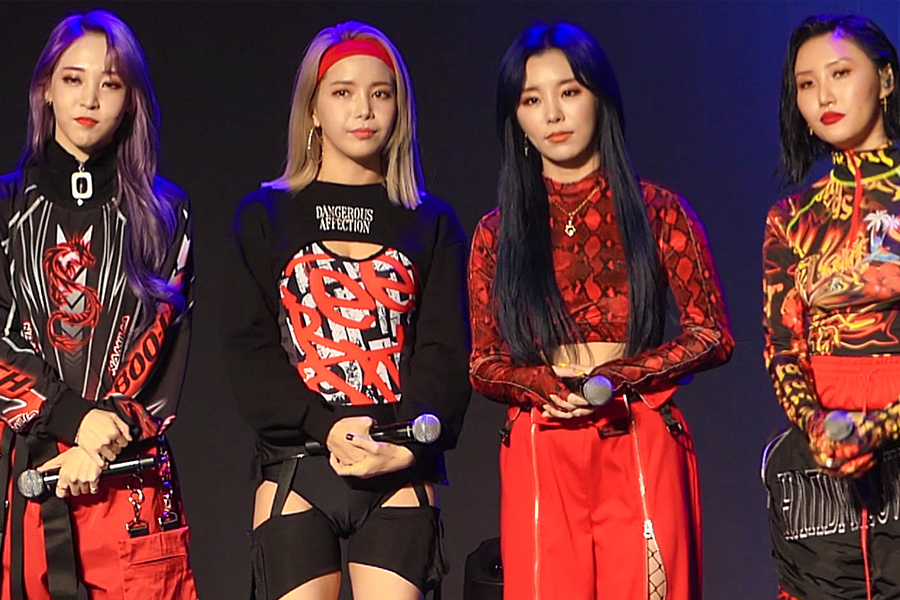 韩国女团MAMAMOO的成员们穿着红色和黑色的华丽舞台服装,在舞台上手持麦克风准备表演。