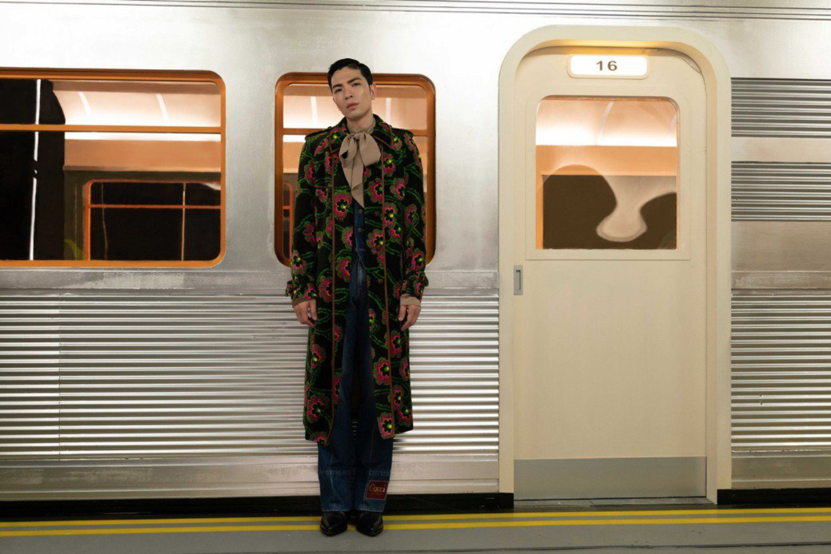 一位身著華麗印花外套和牛仔褲的人物，站立在地鐵車廂門前，衣著獨特時尚，地點設在地鐵站。