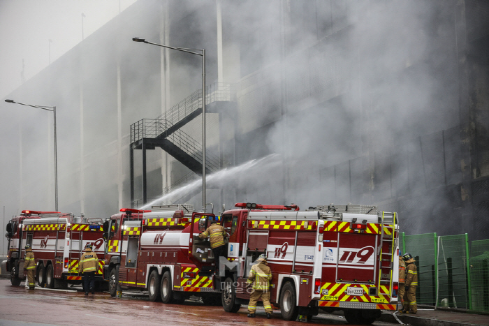 多輛消防車正在倉庫火災現場進行滅火工作，由多位消防員操控水流。