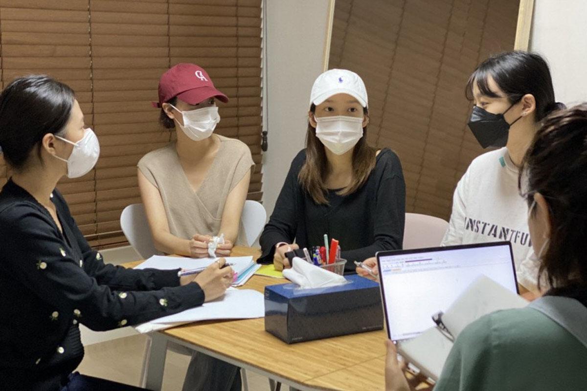 สมาชิกวง APRIL กำลังมีการสัมภาษณ์เพื่อหารือเกี่ยวกับสถานการณ์ในปัจจุบันพร้อมกับปากกาและกระดาษบนโต๊ะ