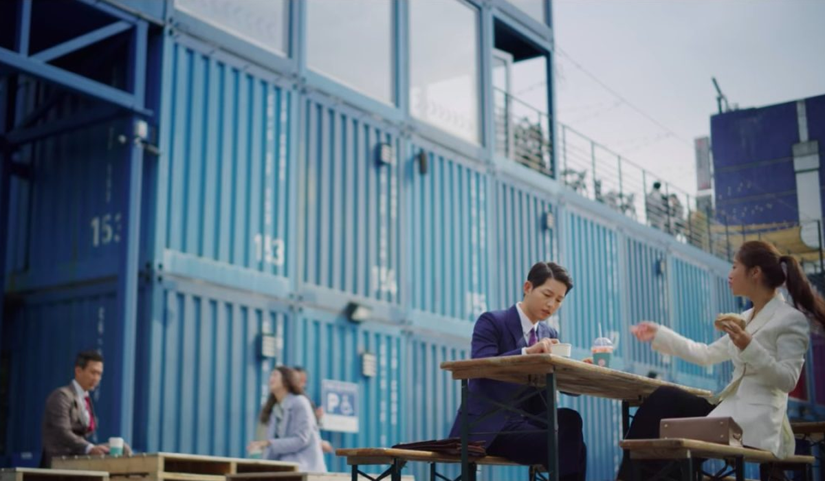 Personaggi seduti a un tavolo esterno presso Common Ground circondato da container blu.