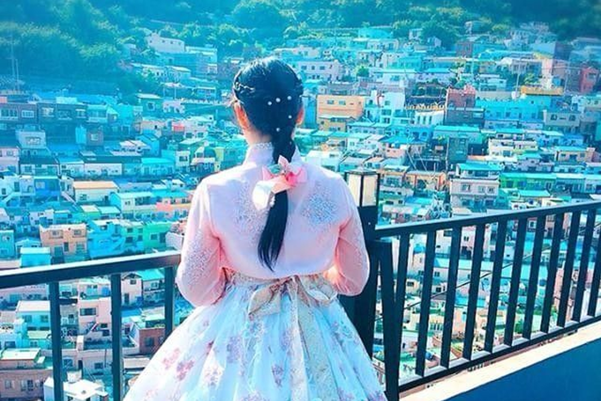釜山甘川文化村觀景台上的女子，她身穿粉嫩色調的韓服，背面面向著五顏六色的房屋，展現韓國傳統服飾與文化景觀的完美融合。