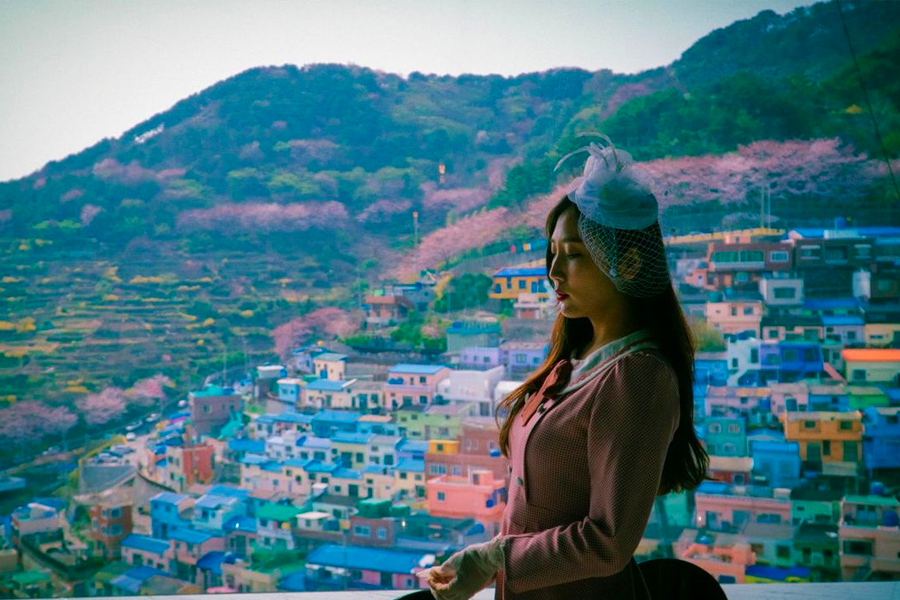 Overlooking colorful houses of Gamcheon Culture Village, a woman in vintage clothing gazes out the window, capturing the scenic beauty.