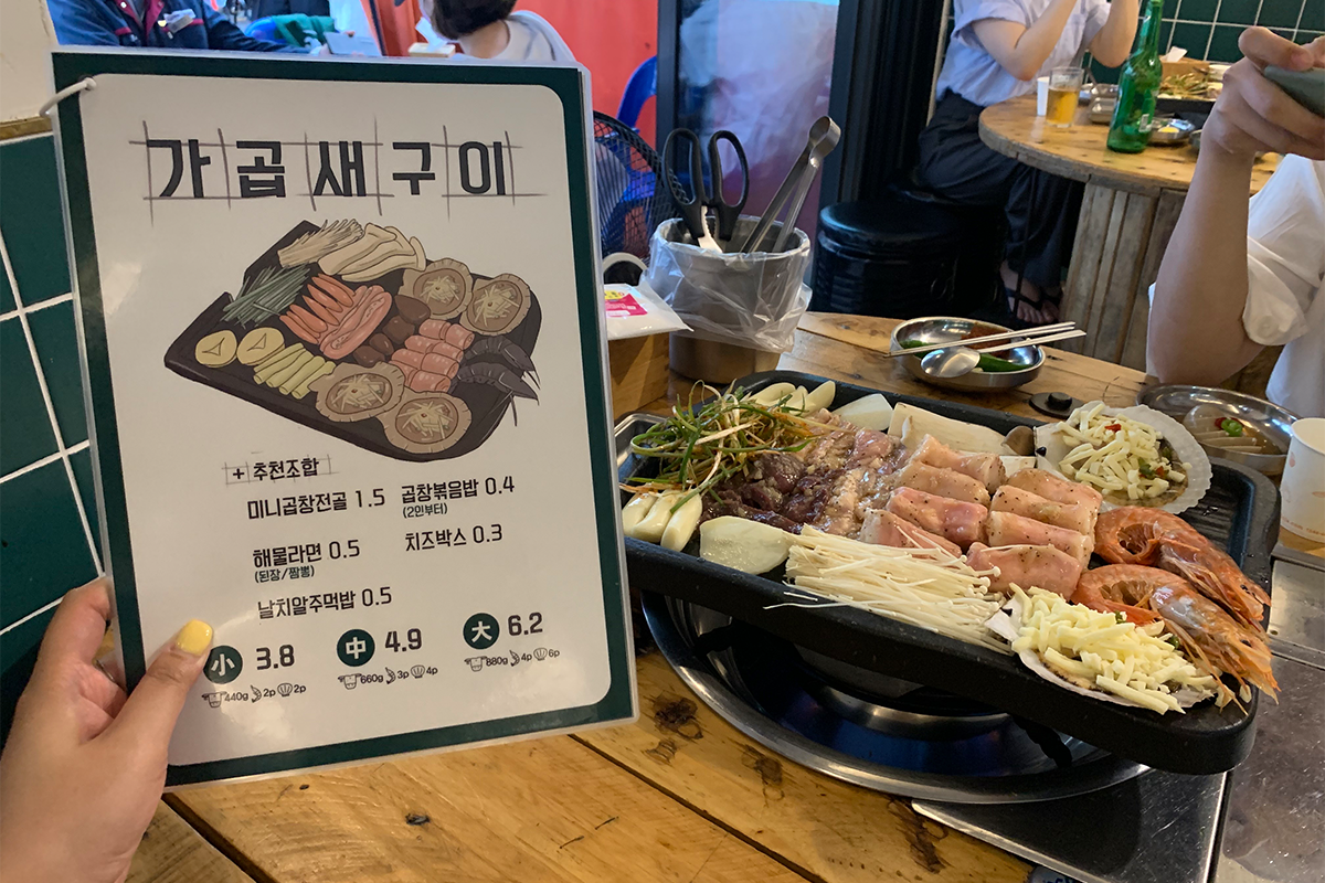 Menu of grilled tripe restaurant in Busan next to grill cooking tripe, seafood, and vegetables on table