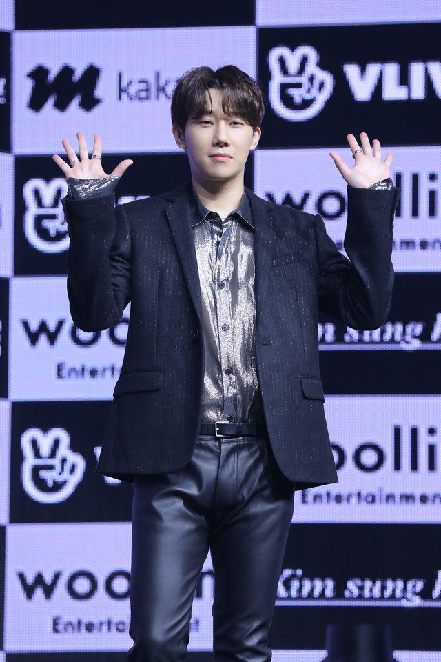 グループINFINITEの元メンバーである韓国の歌手キム・ソンギュが公式イベントでカメラに向かって立っている様子。彼はシンプルな黒いスーツにシルバーのシャツを組み合わせたスタイルを着用しており、背景にはV LIVEやウリムエンターテインメントのロゴが表示。