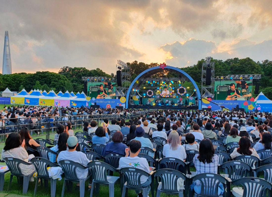 ผู้ชมกำลังนั่งชมคอนเสิร์ตบนเก้าอี้พลาสติกกลางแจ้งที่งานเทศกาลดนตรี Beautiful Mint Life 2021 ที่สวนโอลิมปิก โซล, เกาหลีใต้