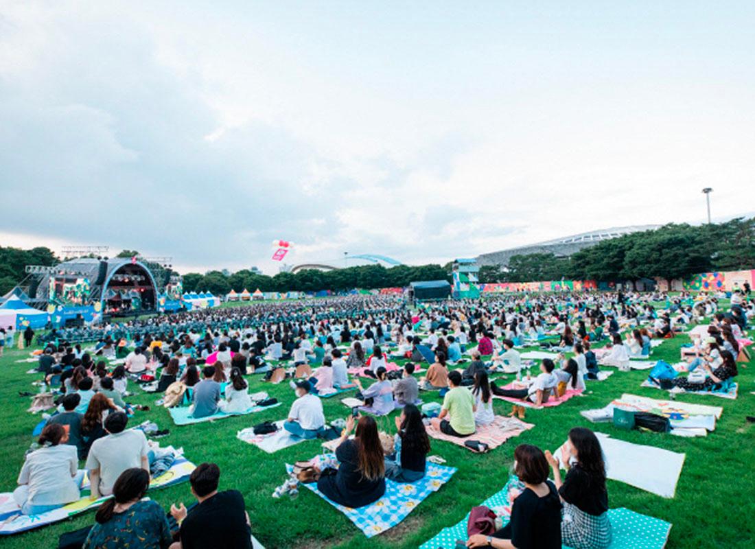 ภาพจากมุมสูงของการแสดงคอนเสิร์ตกลางแจ้งที่สวนโอลิมปิก โซล โดยผู้ชมได้นั่งบนสนามหญ้า ชมการแสดงบนเวทีที่สวยงาม