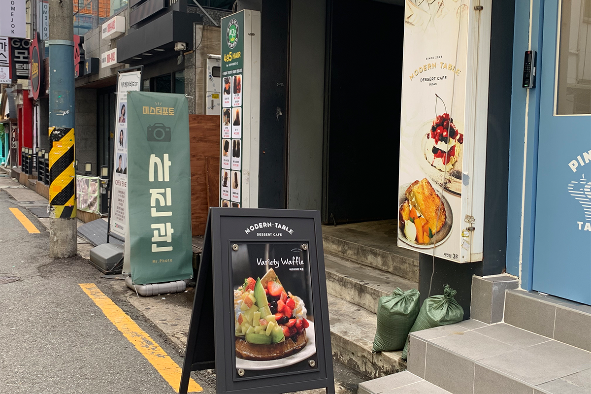 釜山現代桌子咖啡館的入口，外面有一個招牌顯示多種水果鬆餅圖片，旁邊有一個長方形畫畫檔，展示鬆餅和派的圖案。