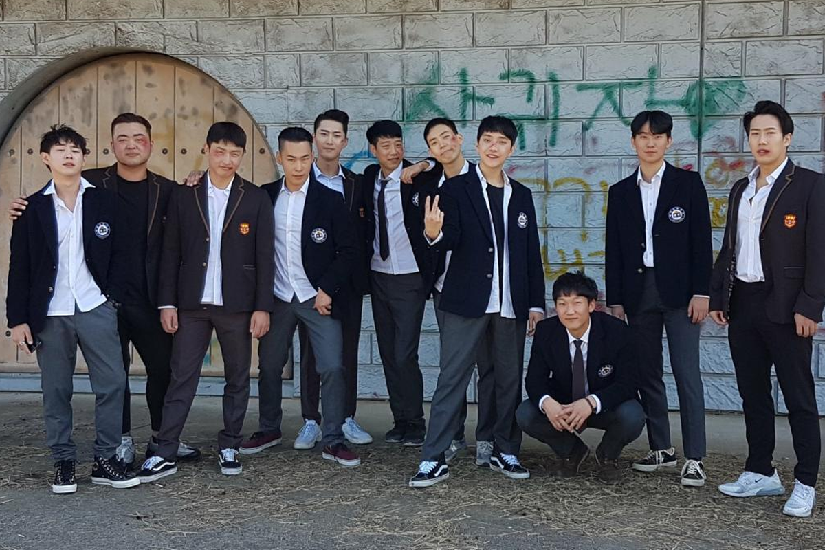 一群穿著校服的青少年在韓國的某個場地合影，背景是以塗鴉牆為主