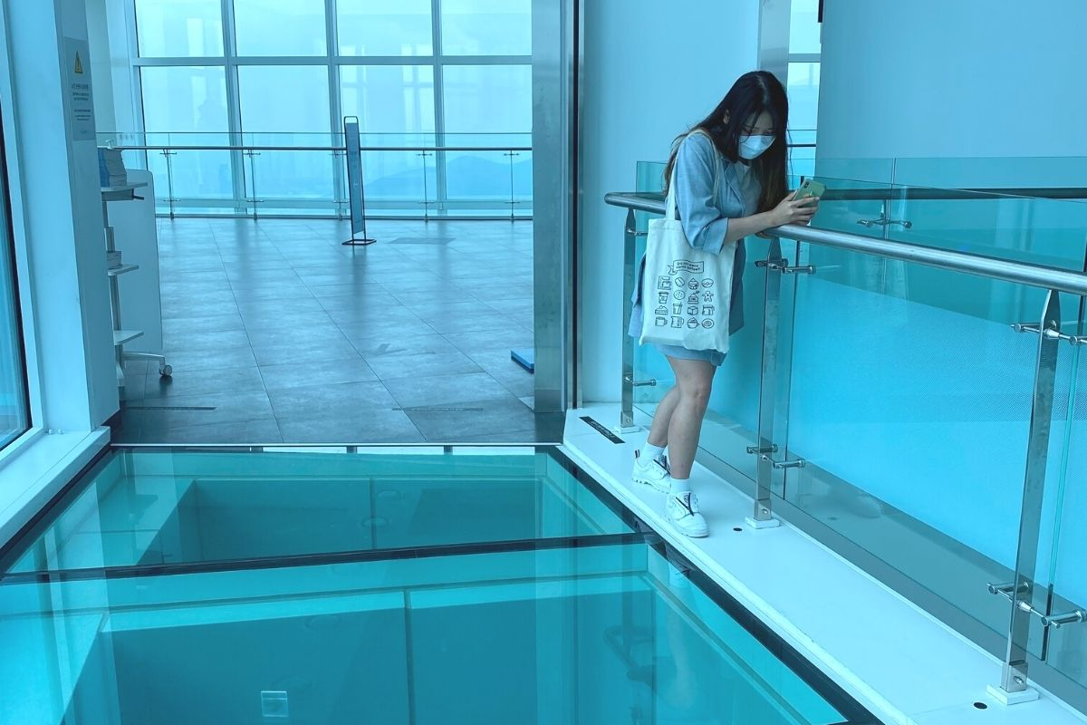 遊客站在透明玻璃走道上，背景是高樓大廈的城市景，視野開闊，是一個可以感受到高空俯瞰美景之地。