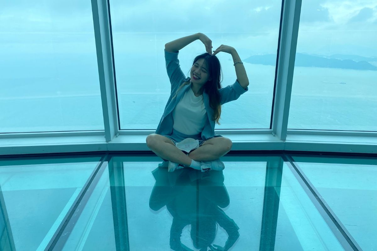 遊客坐在透明地板上，用雙手比出心形，背景是寬廣的海景，顯示出這裡是觀光愛好者的理想打卡地點。