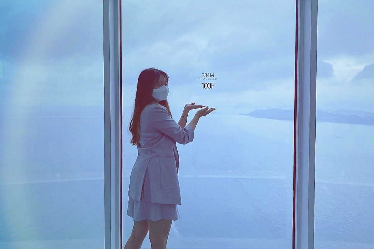 室內觀景窗前，一位遊客在欣賞窗外的釜山全景，窗上標記著384米和100樓的高度顯示，使人感受到高處的壯觀。
