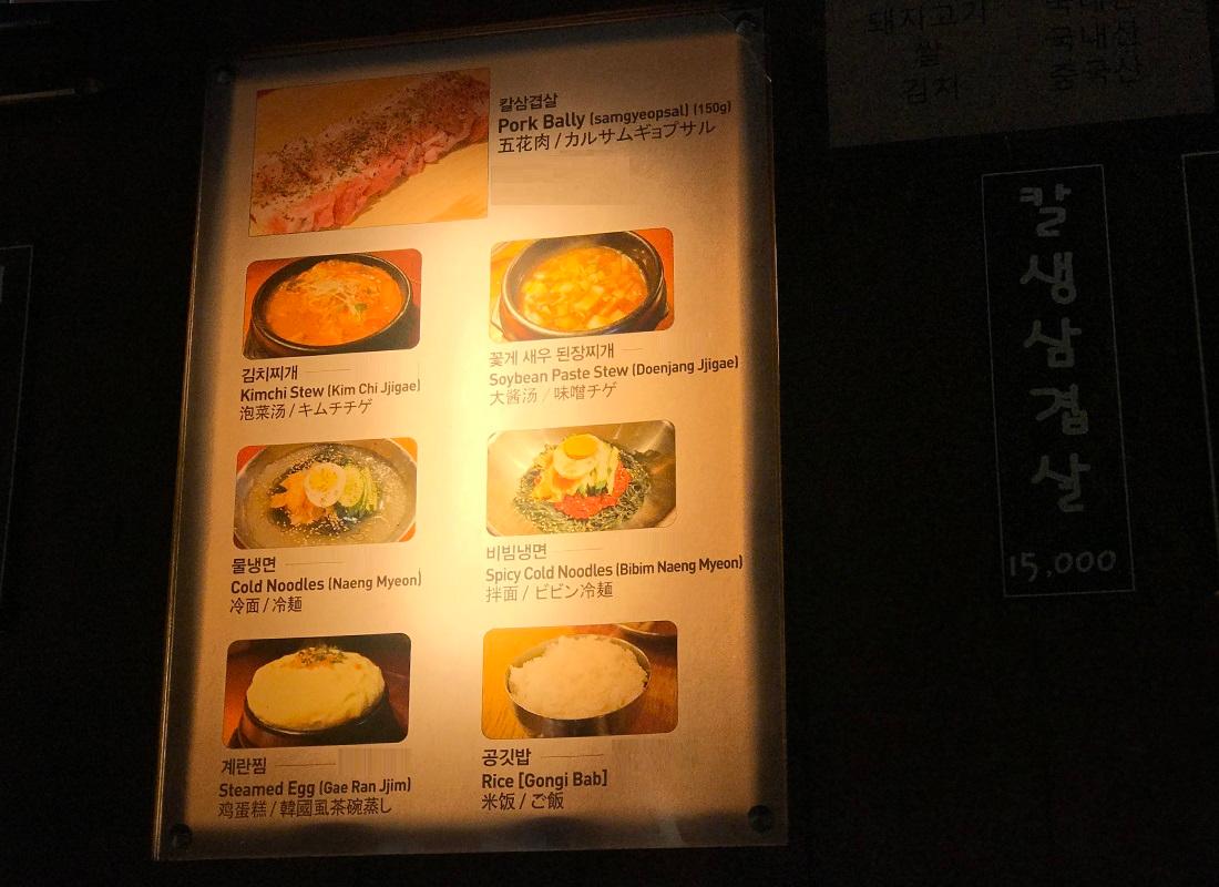 店内の多言語メニュー表示：韓国料理のカルサムギョプサル、キムチチゲ、冷麺、ビビン冷麺、韓国式蒸し卵、白米などのメニューが書かれた光るボード