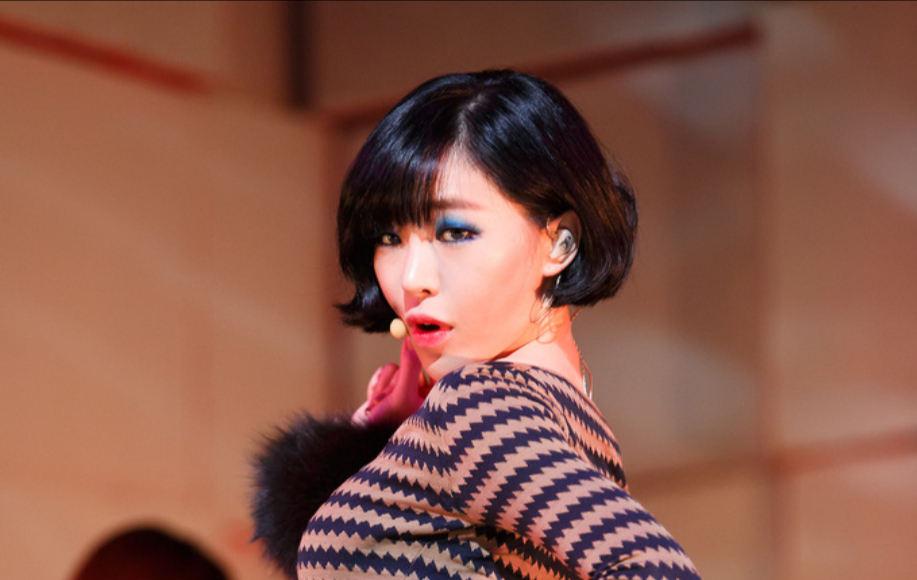 茶色のジグザグ模様のドレスを着たショートカットの女性、背景には赤いライトで照らされたステージが見える。