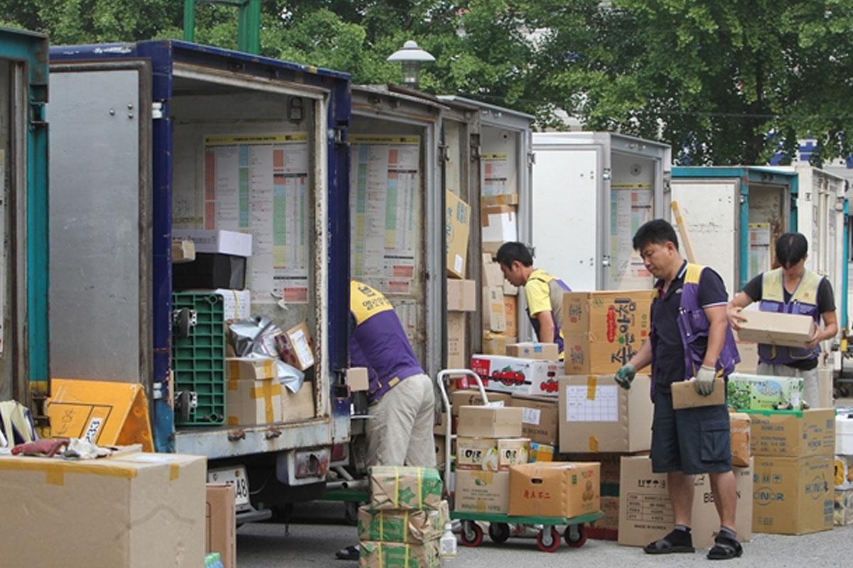 พนักงานสวมเสื้อสีม่วงและเหลืองกำลังขนของจากรถบรรทุกในพื้นที่การจัดส่งสินค้าของเกาหลีใต้