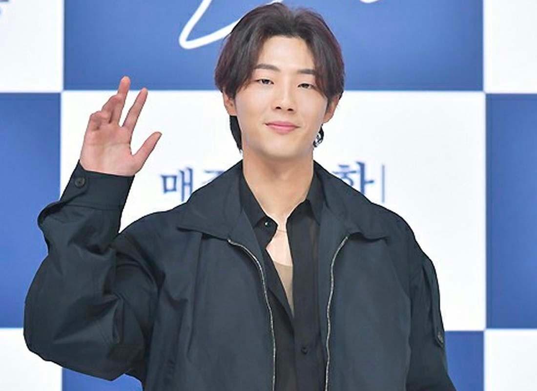 ภาพนักแสดงเกาหลีคนหนึ่งสวมสูทและเสื้อสีน้ำเงิน ด้านหลังเป็นป้ายสีน้ำเงินและขาว