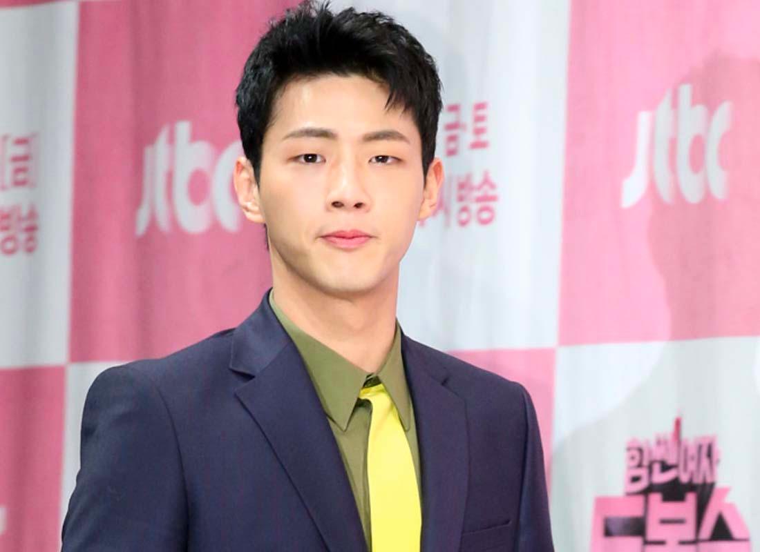 นักแสดงชายเกาหลีสวมสูทสีเทาคู่กับเนคไทสีเหลือง หน้าป้ายเหตุการณ์สีชมพู