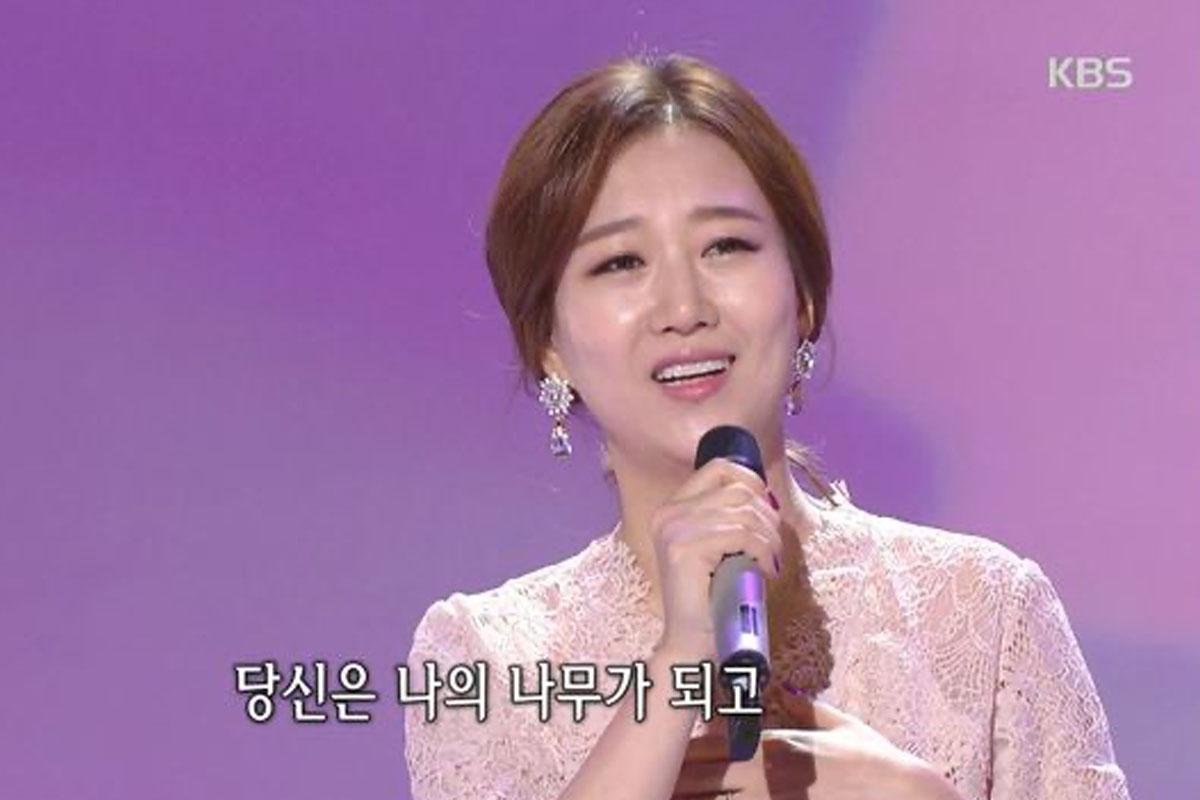 นักร้องยืนอยู่บนเวทีถือไมโครโฟน พร้อมด้วยเนื้อเพลงที่ปรากฏบนหน้าจอในภาษาเกาหลี