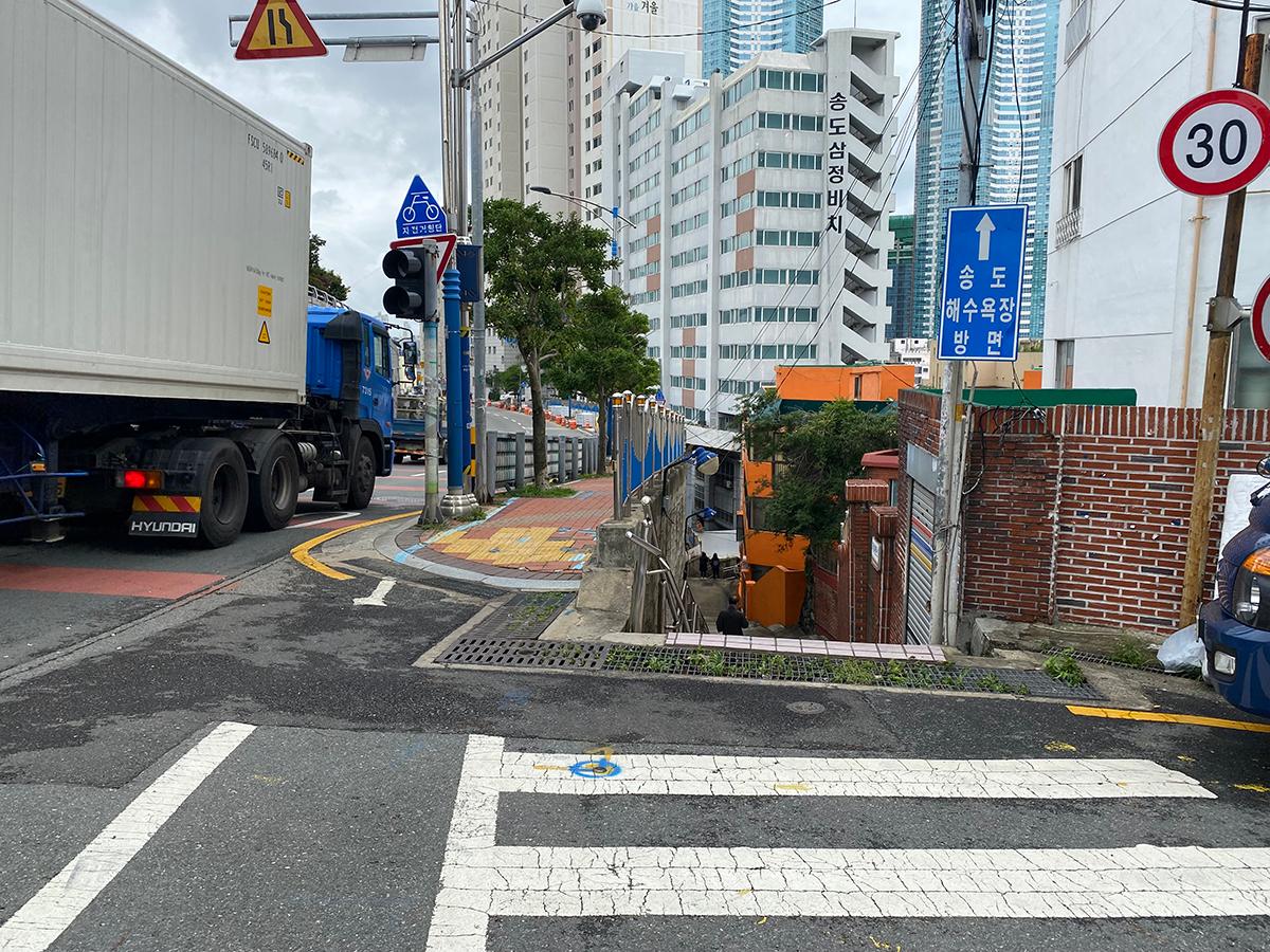 A Busan street with a sign for Songdo Beach, showing a nearby bus and tall buildings in the background.