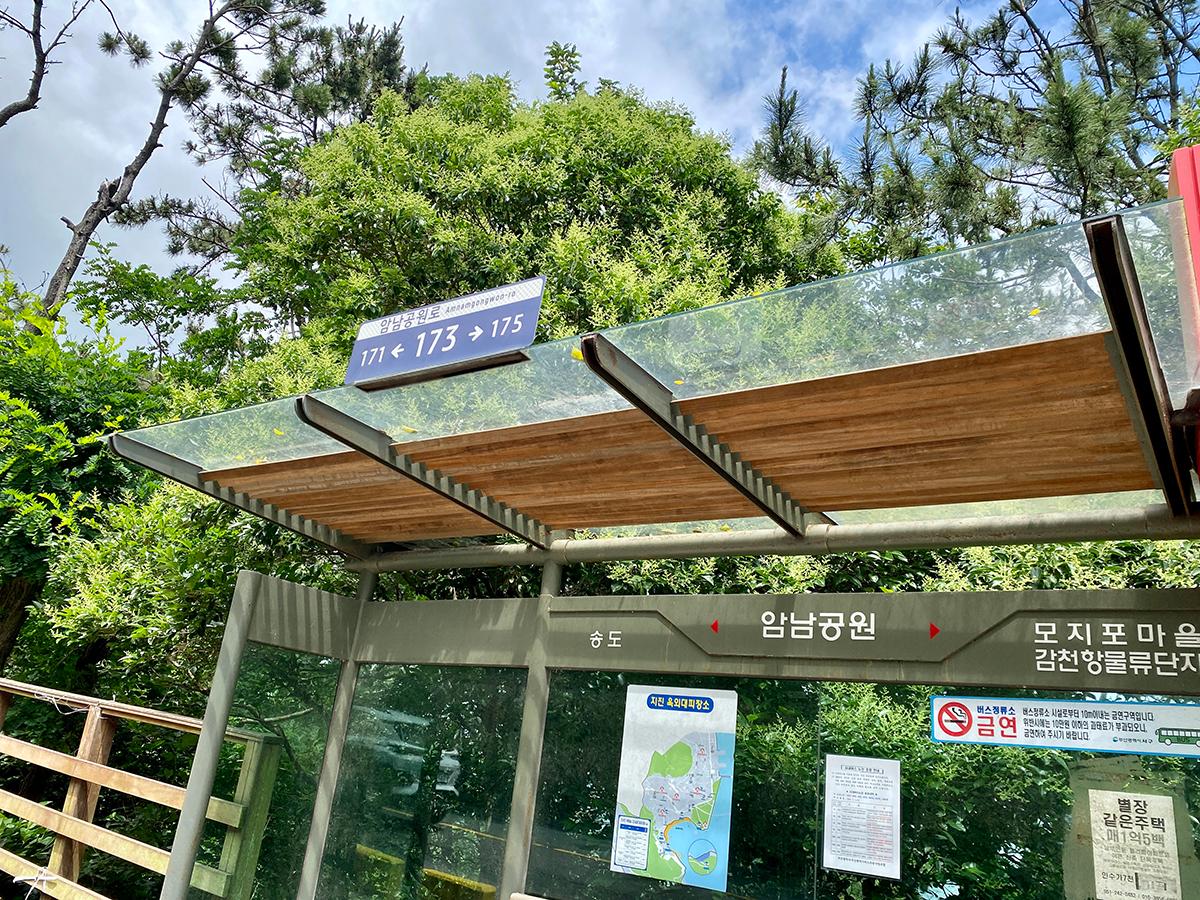 Bus stop shelter in Amnam Park, surrounded by green trees with signs indicating bus numbers.