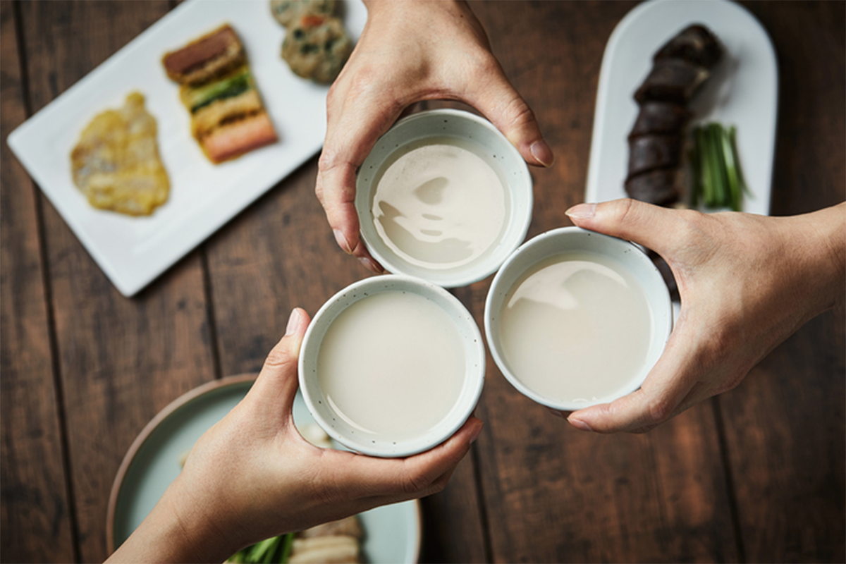 三隻手持著裝有米酒的碗，桌面上放有韓國食物，説明為何米酒通常使用較大容器。