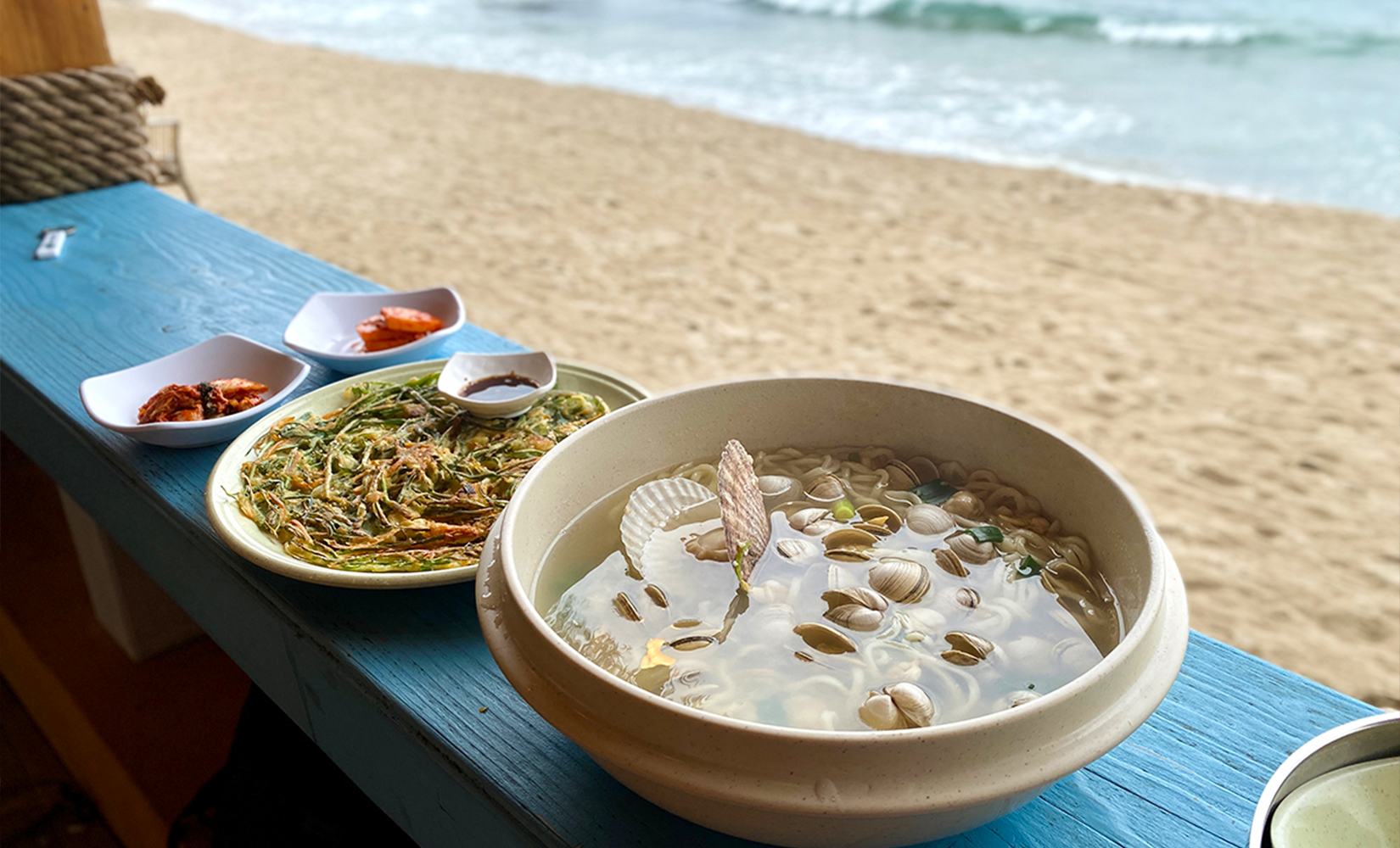 From back to front, two kimchi dishes, a chive pancake and kalguksu (seafood noodles) on a blue table in Busan