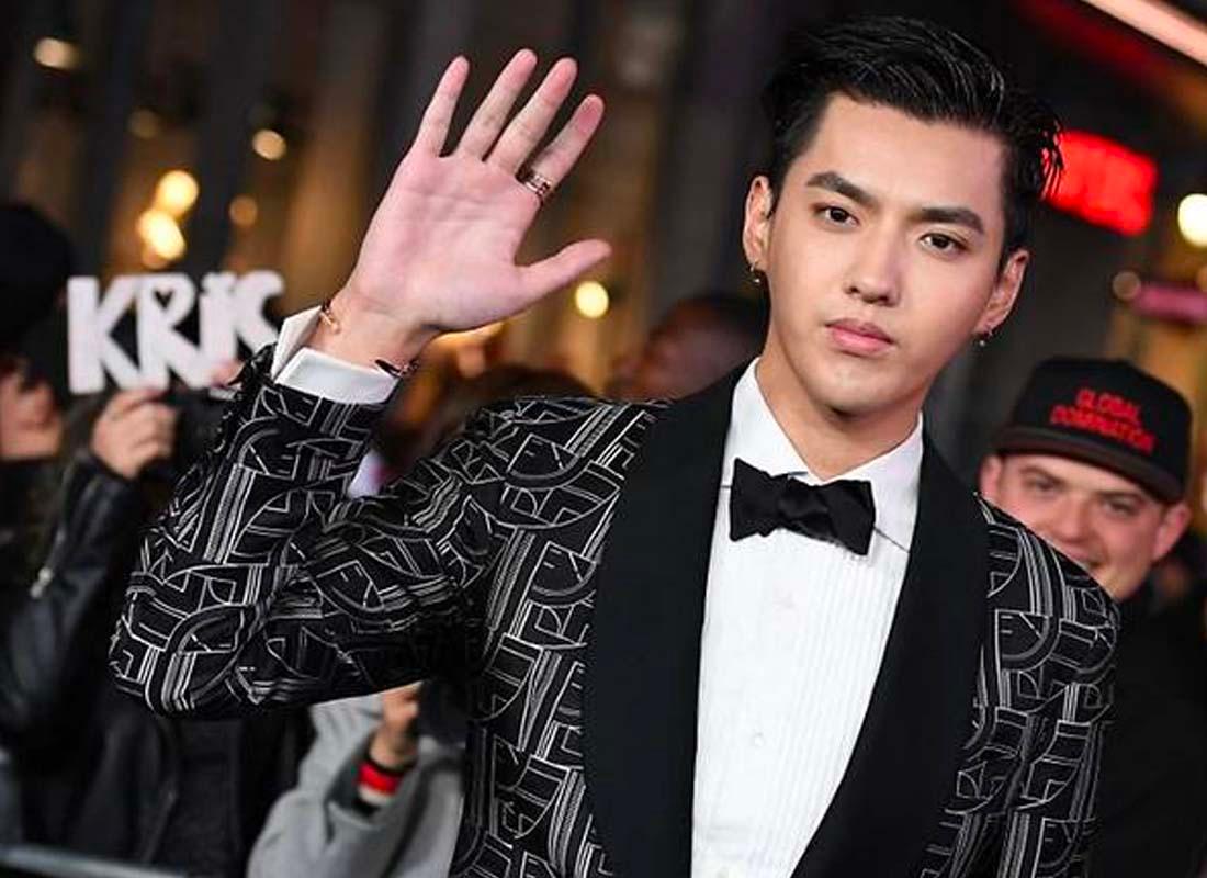 บุคคลสวมชุดทักซิโด้สีดำยืนอยู่ในงานอีเวนท์ที่มีผู้คนมองเห็นอยู่ในพื้นหลังพร้อมป้ายชื่อคริสทะเลาะกันเบื้องหลัง