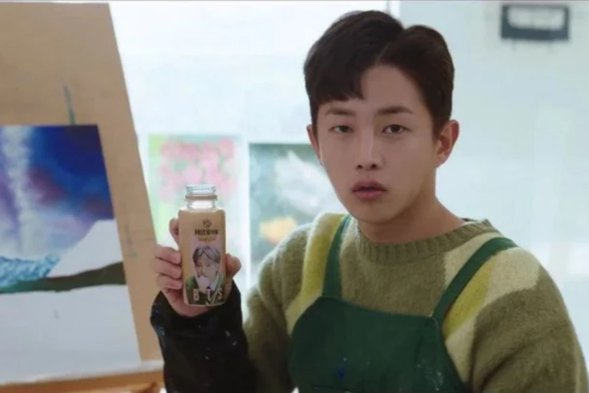 一個人舉著一瓶熱可可飲品，背景中有一些繪畫作品。